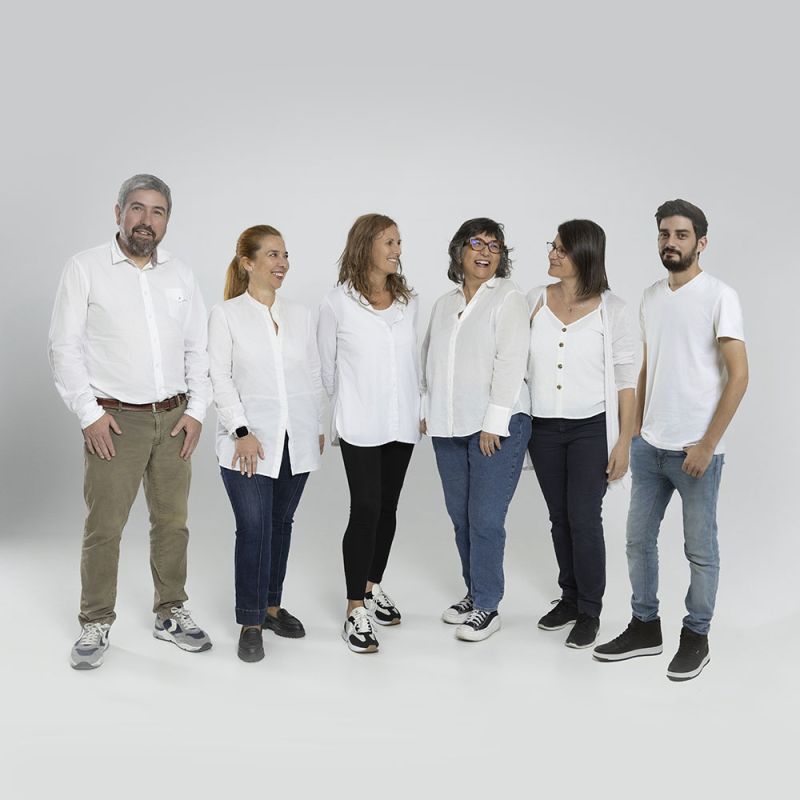A woman with long brown hair wearing a white sleeveless top is posed resting her arms and head over a white table. Positioned against her arm is a sleek, modern massage device in a dark green color with a gold accent.