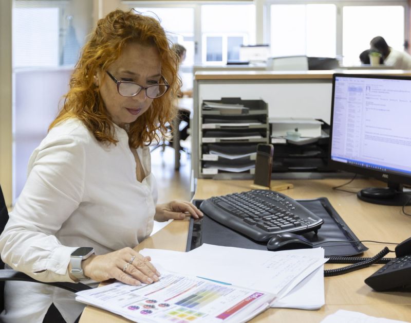 A woman with long brown hair wearing a white sleeveless top is posed resting her arms and head over a white table. Positioned against her arm is a sleek, modern massage device in a dark green color with a gold accent.